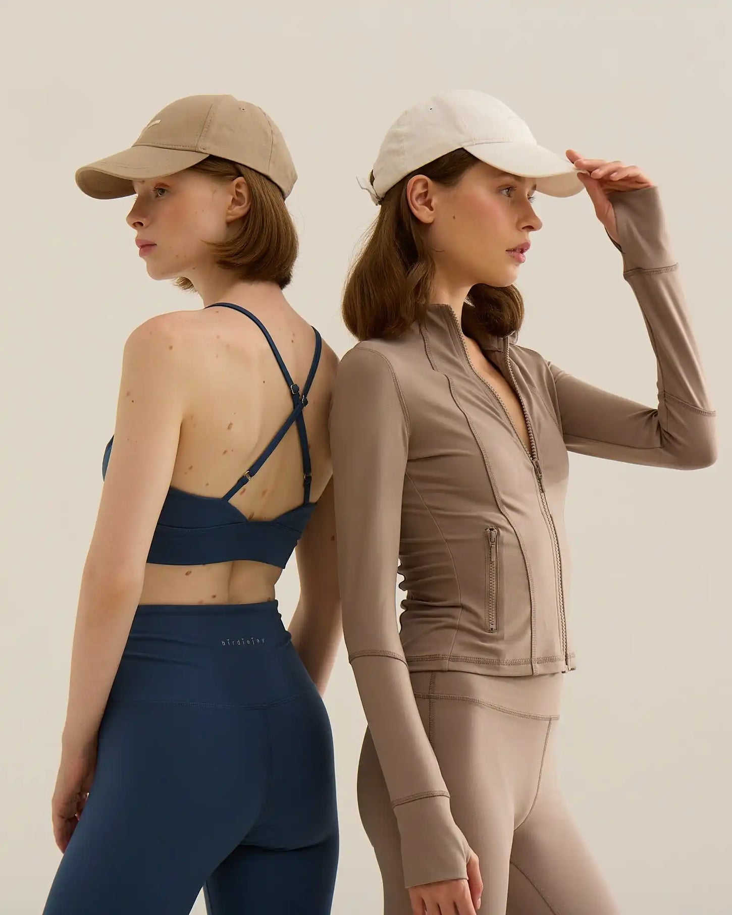 İki kadın spor kıyafetleri içinde, biri mavi sporcu sütyeni ve tayt, diğer ise taupe fermuarlı ceket ve tayt giymiş, ikisi de şapka takmış.